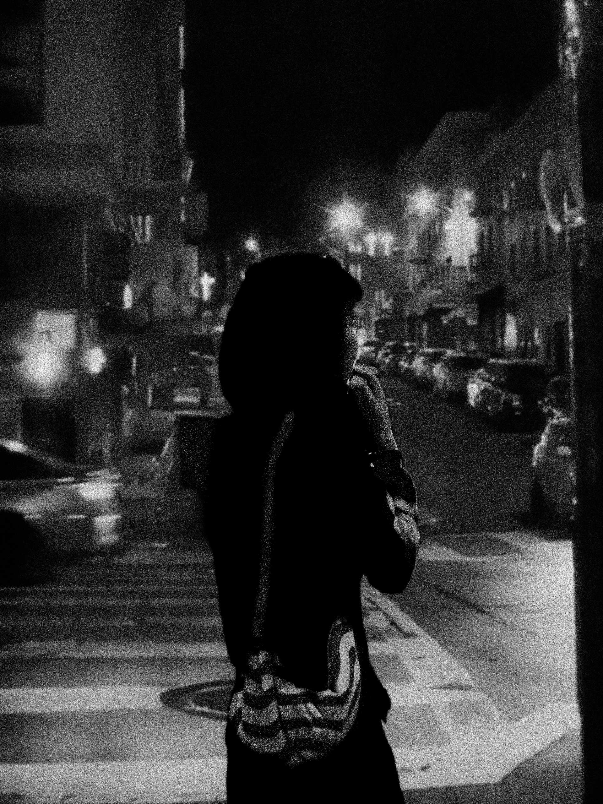 A grainy black-and-white portrait of a person silhouetted on a city sidewalk at night, with parked cars and streetlights behind them.