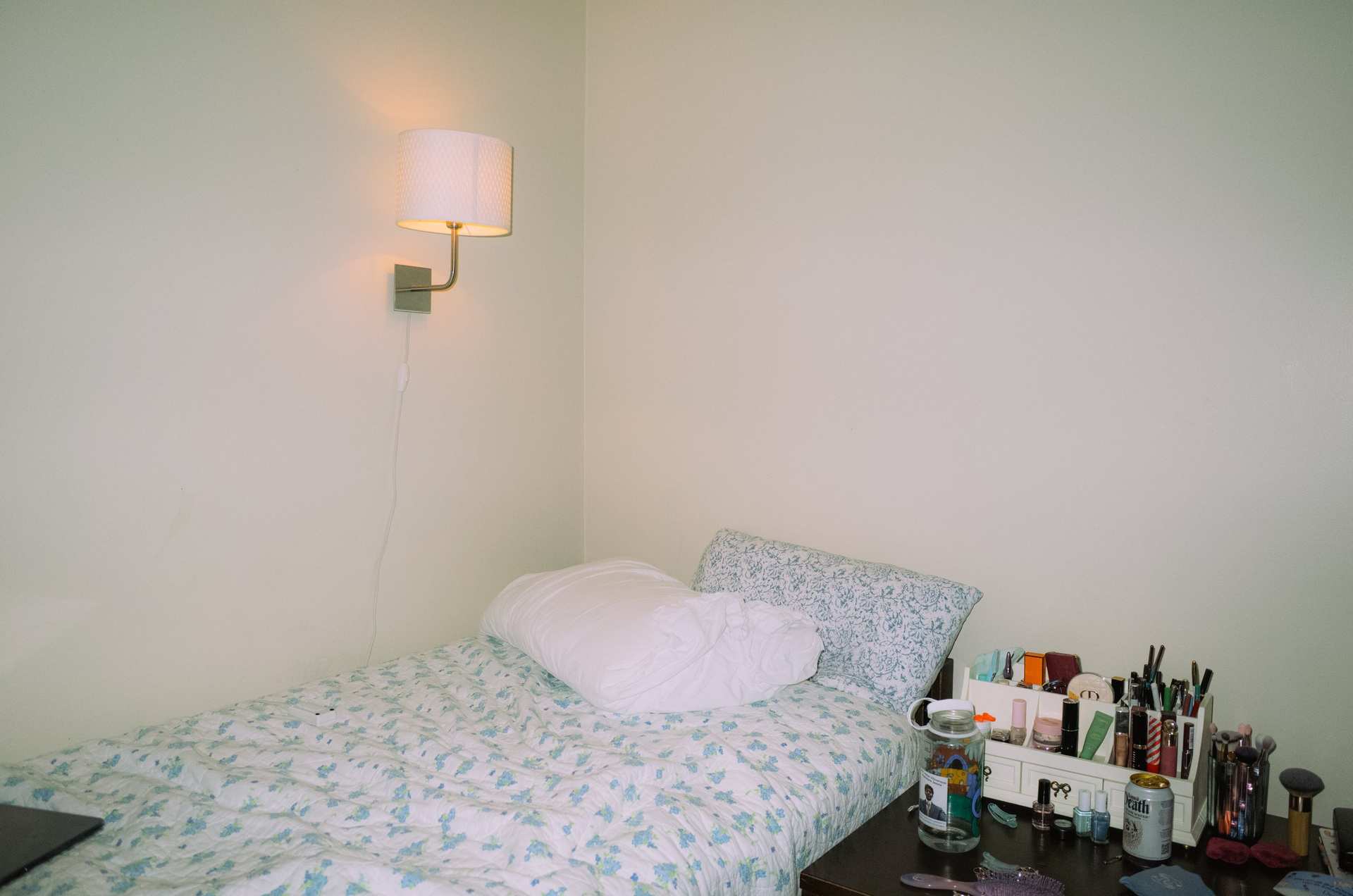 A bed with floral bedding beneath a glowing wall lamp, beside a collection of makeup brushes.