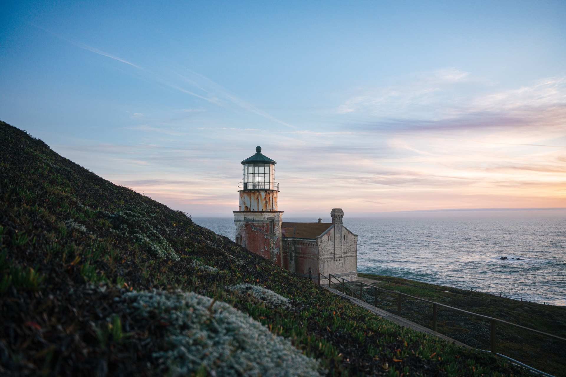 A lighthouse on a coastal hillside overlooking the ocean at sunset.