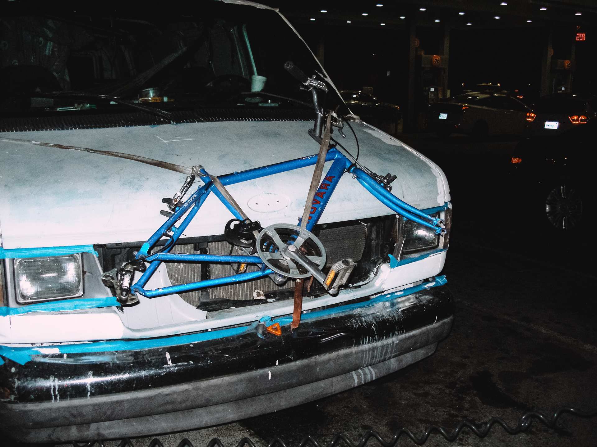 A blue bicycle frame strapped across the front of a weathered white van at a gas station at night.