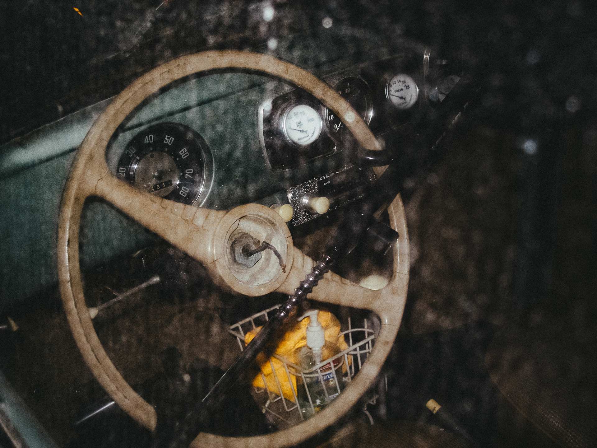 A wooden steering wheel and dashboard gauges seen through the reflective windshield of a vintage vehicle.