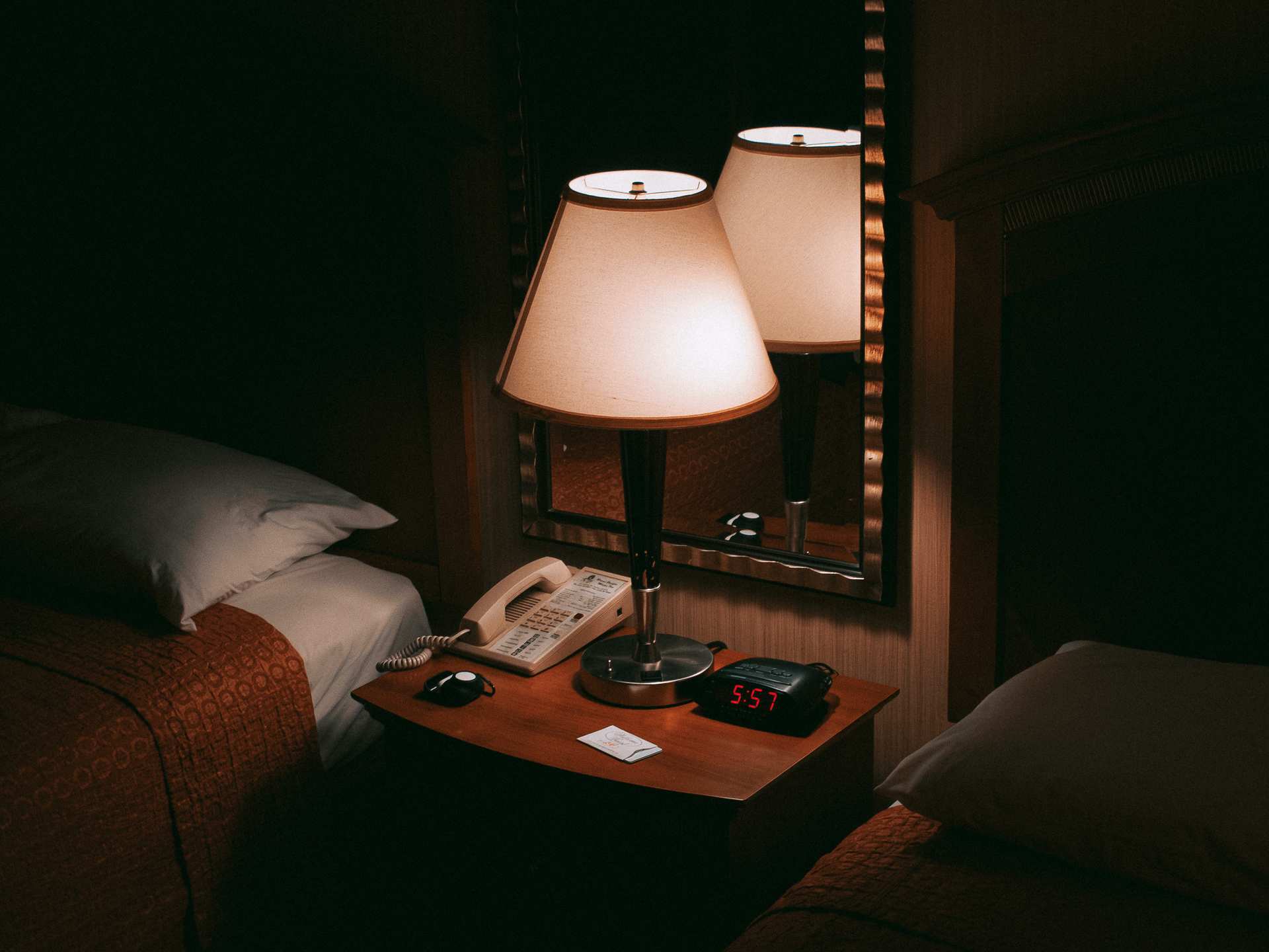 A glowing bedside lamp reflected in a mirror above a telephone and digital clock between two hotel beds.