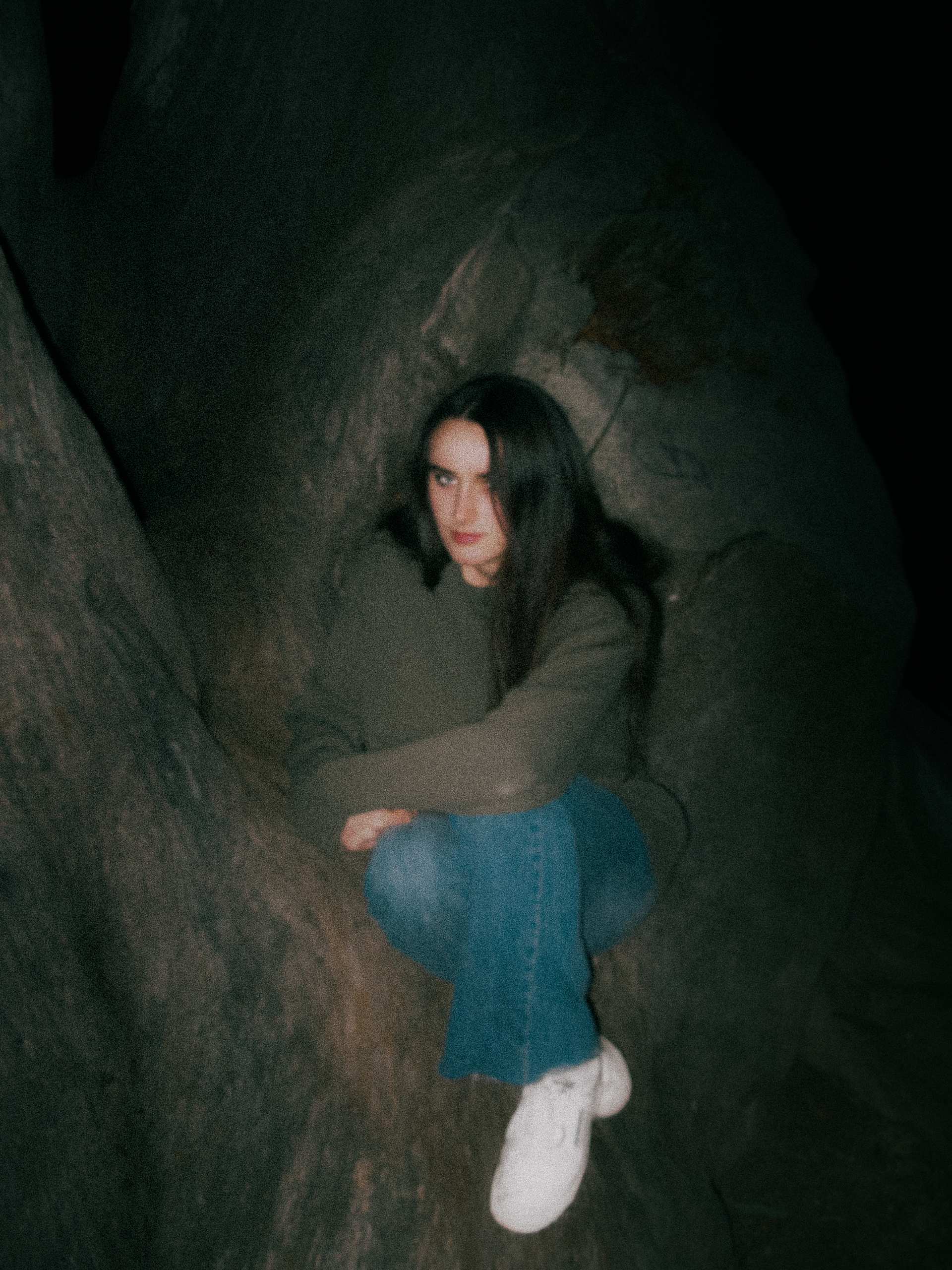 A woman in a green sweater and blue jeans seated inside the hollow of a large tree.