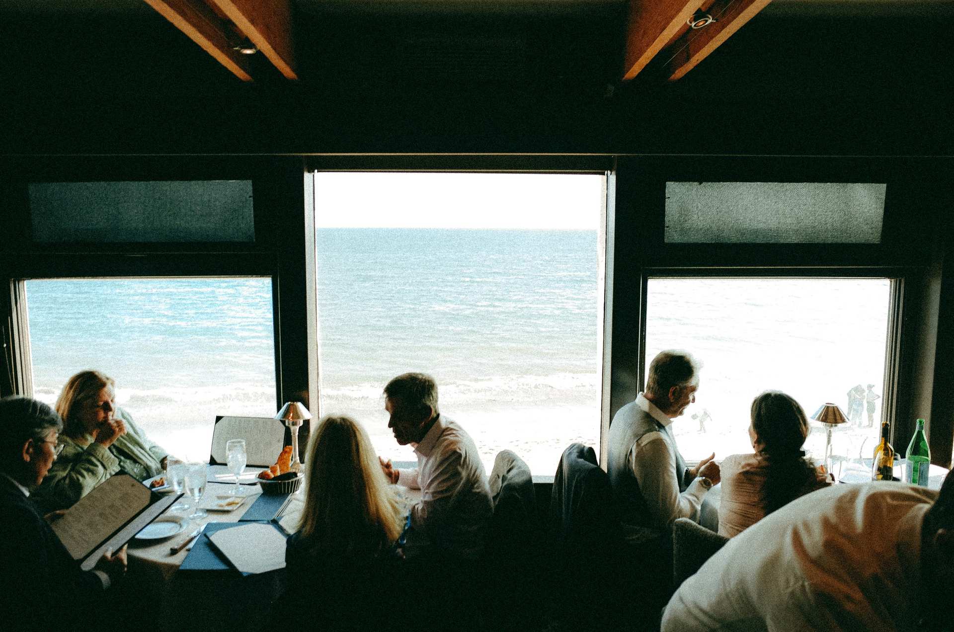 Diners silhouetted in a restaurant, with three bright windows looking out over the ocean.