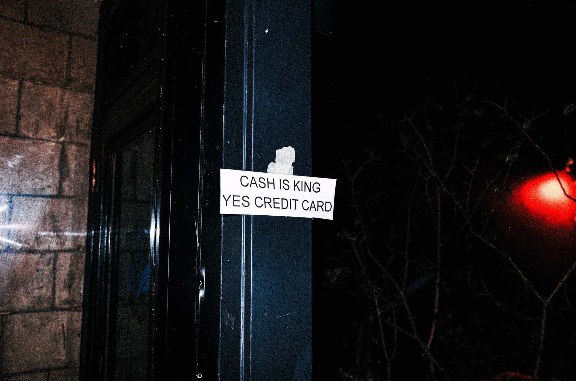 A taped “Cash is king, yes credit card” sign on a dark blue door, with a red light reflected at the right.