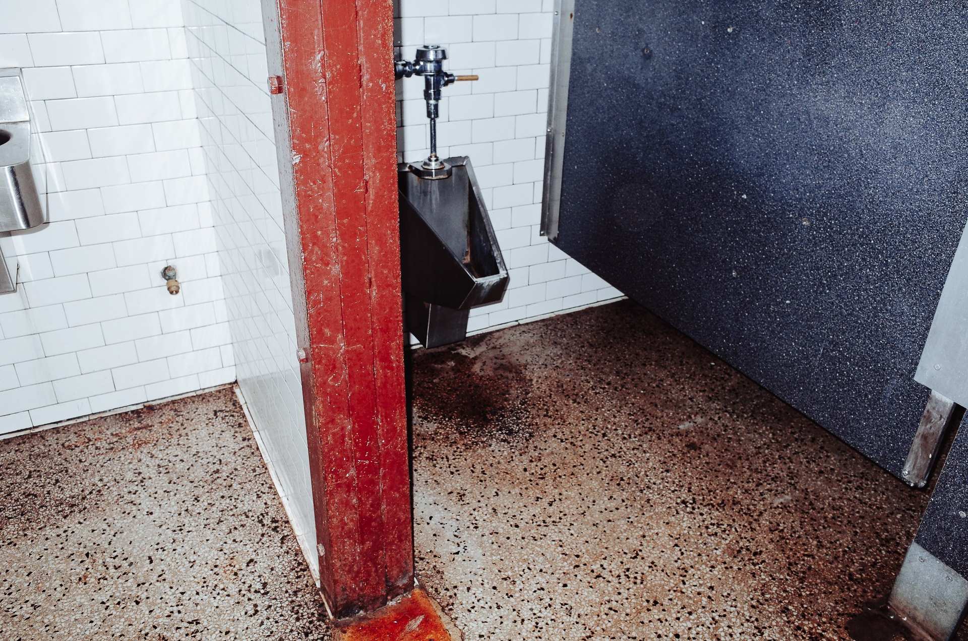 A red door frame dividing white tiles and a dark blue restroom stall, above a worn terrazzo floor.
