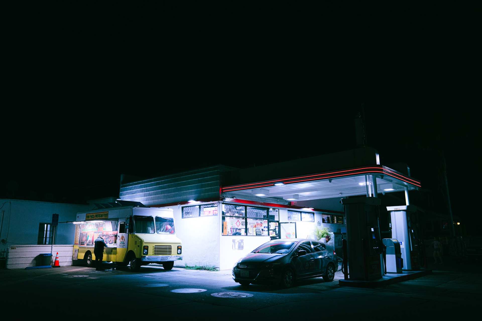 A yellow Volkswagen van and a dark car at a brightly illuminated gas station at night.